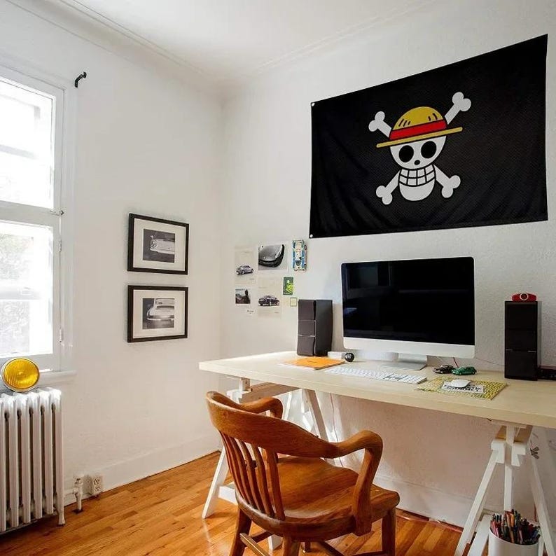 May include: A black flag with a white skull and crossbones design, wearing a yellow and red straw hat, hangs on a white wall. The flag is above a wooden desk with a computer, speakers, and a wooden chair. The room has a window and hardwood floors.