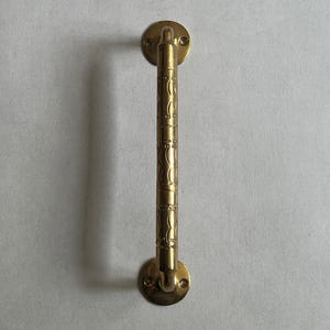May include: A gold-colored door pull with a decorative, textured design. The pull has a cylindrical handle with repeating patterns and circular mounting plates at each end. The hardware is mounted on a light gray surface.