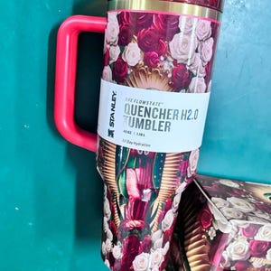 May include: A red and floral Stanley Quencher H2.0 Tumbler with a handle. The tumbler features a design of red and white roses and the text "QUENCHER H2.0 TUMBLER". It holds 40 oz.