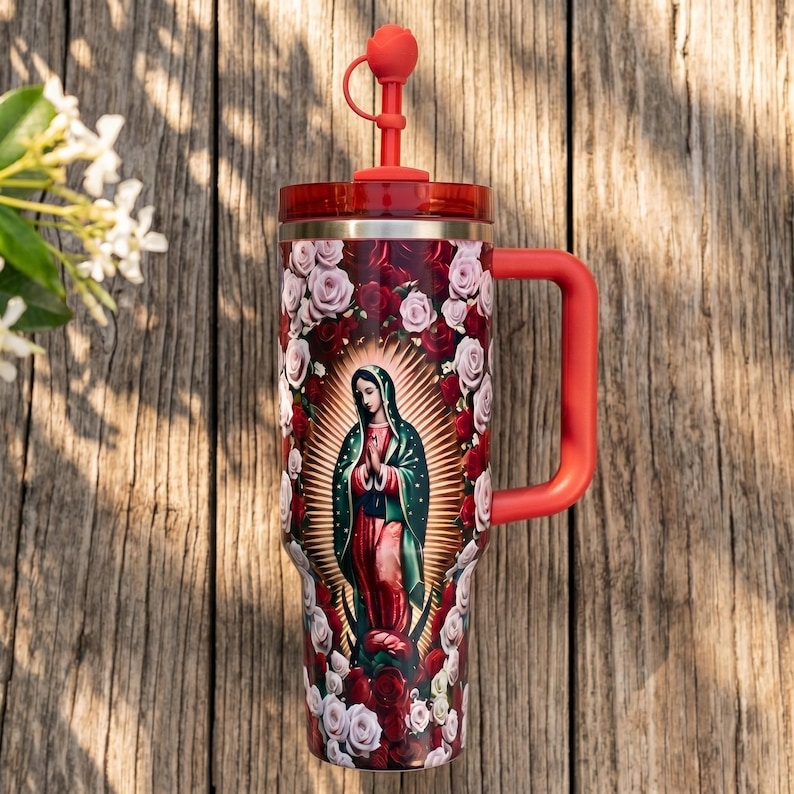 May include: A large red and stainless steel tumbler featuring an image of Our Lady of Guadalupe surrounded by roses. The tumbler has a red handle, lid, and straw with a heart-shaped top. The background is a wooden surface.