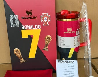 May include: A red and black Stanley travel mug with a matching box featuring the name "RONALDO" and the number "7". The box also displays the Stanley logo, a Portuguese crest, and two golden trophy figures.