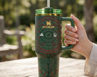 May include: Green and red patterned Stanley tumbler with a handle and straw. The tumbler features the Stanley logo, Adidas logo, and other graphics. The tumbler is held by a person's hand.