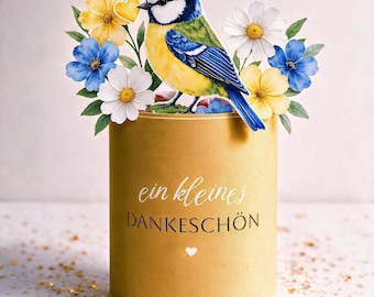 May include: A cylindrical gold container featuring the text "ein kleines DANKESCHÖN" and a small heart icon. A blue, yellow, and white bird illustration sits atop the container, surrounded by yellow and white daisies and blue flowers.