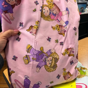 May include: Pink fabric with a repeating pattern of cartoon dolls, butterflies, and flower pots. The dolls are wearing purple and pink outfits. The butterflies are in various colors. The fabric is folded and held by a hand.