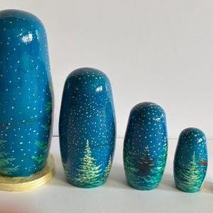 Puede incluir: Un conjunto de cinco mu&ntilde;ecas rusas, cada una pintada con un dise&ntilde;o de cielo nocturno azul oscuro y detalles de estrellas blancas. Las mu&ntilde;ecas presentan una escena de bosque con &aacute;rboles verdes y una base dorada.