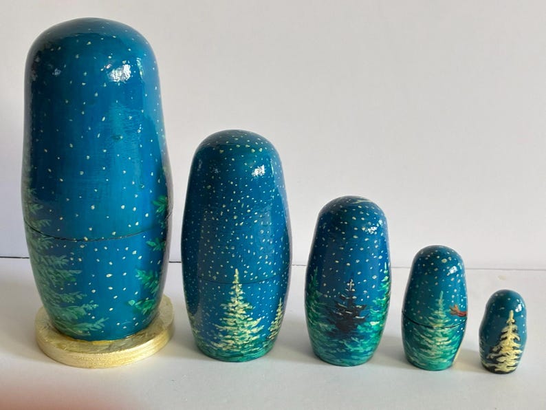 Puede incluir: Un conjunto de cinco mu&ntilde;ecas rusas, cada una pintada con un dise&ntilde;o de cielo nocturno azul oscuro y detalles de estrellas blancas. Las mu&ntilde;ecas presentan una escena de bosque con &aacute;rboles verdes y una base dorada.