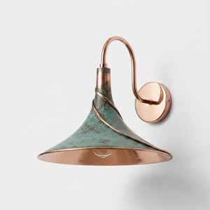 May include: A wall-mounted lamp featuring a flared, conical shade with a weathered, teal-green patina finish and a polished copper trim. The fixture includes a curved copper arm and a circular wall plate, with a single light bulb visible inside.