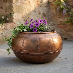 May include: A round, hammered copper planter featuring a riveted panel design. The planter is filled with vibrant purple petunia flowers, a small green succulent, and trailing ivy vines, creating a contrast between the metallic finish and plants.