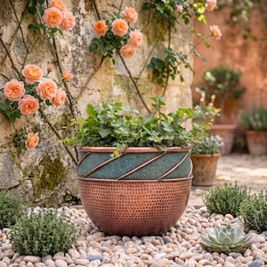 Op de afbeelding: Een ronde plantenbak met een basis van gehamerd koper en een blauwgroen bovengedeelte met diagonale koperen banden. De plantenbak bevat een groene bladplant en staat op een bed van lichtgekleurde kiezels bij een stenen muur met rozen.