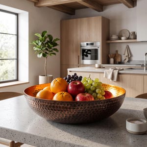 Puede incluir: Un cuenco grande y redondo de metal martillado con un exterior de bronce oscuro y un interior de oro pulido. El cuenco está lleno de fruta fresca, incluyendo naranjas brillantes, manzanas de color rojo intenso y uvas verdes y moradas.