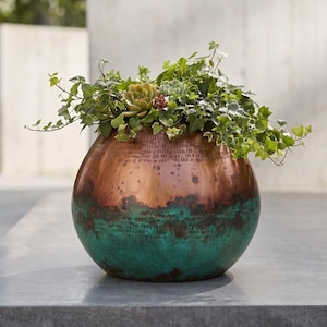 May include: A spherical metal planter featuring a polished copper upper section and a textured, oxidized teal green lower section. The planter is filled with a lush arrangement of green ivy vines and several small, fleshy succulent plants.