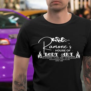 Puede incluir: Camiseta negra con un diseño blanco que dice "Ramone's House of Body Art". El diseño incluye llamas y el texto "Pin Striping - Flame Jobs - Air Brushing". La camiseta es de cuello redondo.