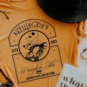 Puede incluir: Camiseta de color amarillo mostaza con un dise&ntilde;o negro de Jack Skellington y Zero de Pesadilla antes de Navidad, con el texto "SKELLINGTON'S PUMPKIN ALE BREWED IN HALLOWEEN TOWN". Tambi&eacute;n se ven un sombrero negro, una c&aacute;mara y un libro.