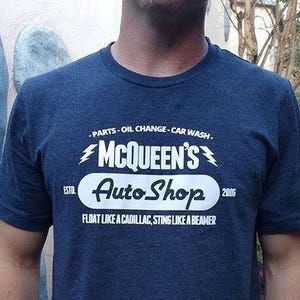 Puede incluir: Camiseta azul marino con texto blanco. La camiseta presenta el texto "MCQUEEN'S Auto Shop" con el lema "FLOAT LIKE A CADILLAC, STING LIKE A BEAMER". También incluye el texto "PARTS - OIL CHANGE - CAR WASH".