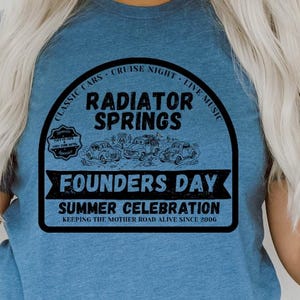 Puede incluir: Una camiseta azul jaspeado de manga corta con un estampado gráfico negro que dice Radiator Springs Founders Day Summer Celebration, Classic Cars, Cruise Night, Live Music y Keeping The Mother Road Alive Since 2006 con ilustraciones.