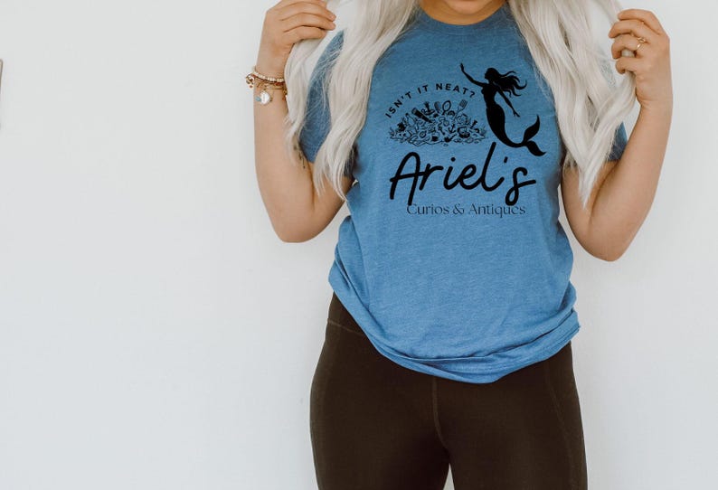 May include: A blue t-shirt with the text "Isn't it neat?" above "Ariel's Curios & Antiques". A black mermaid graphic is on the right side of the shirt. The shirt is worn by a person with blonde hair and dark brown leggings.