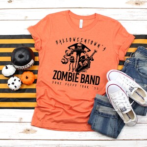 May include: Orange t-shirt with a black graphic of a zombie band, jeans, and white sneakers. The shirt reads "Halloweentown's Zombie Band Done Pappy Tour '95". Decorative pumpkins and a black and orange striped runner complete the Halloween theme.