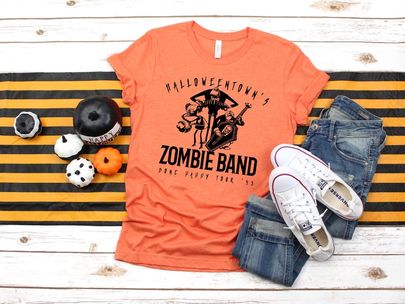May include: Orange t-shirt with a black graphic of a zombie band, jeans, and white sneakers. The shirt reads "Halloweentown's Zombie Band Done Pappy Tour '95". Decorative pumpkins and a black and orange striped runner complete the Halloween theme.