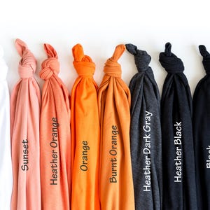 May include: A collection of folded t-shirts in various colors, including white, sunset, heather orange, orange, burnt orange, heather dark gray, heather black, and black. Each shirt has a label with its color name.