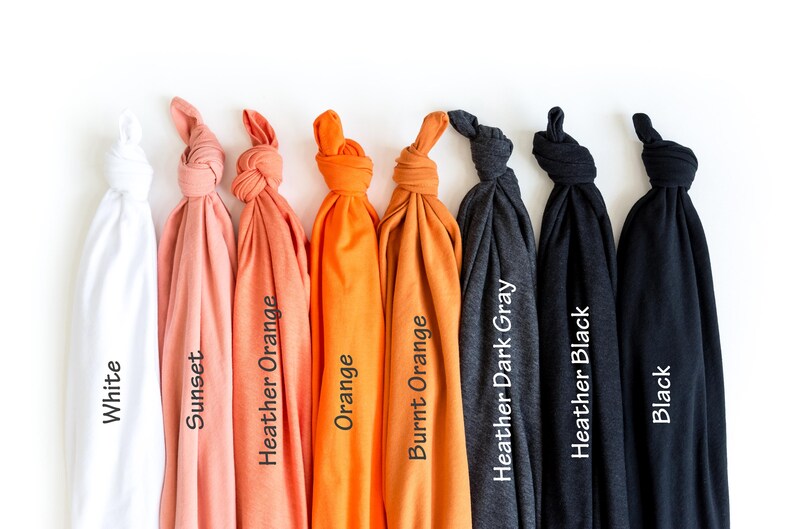 May include: A collection of folded t-shirts in various colors, including white, sunset, heather orange, orange, burnt orange, heather dark gray, heather black, and black. Each shirt has a label with its color name.