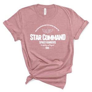 Op de afbeelding: Een stoffig roze t-shirt met de witte tekst "STAR COMMAND". De tekst "SPACE RANGERS" en "to infinity and beyond" staan er ook op. Het t-shirt heeft ook het jaartal "1995" erop staan.