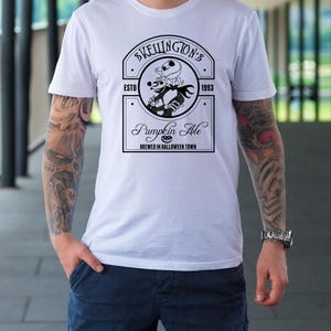Puede incluir: Camiseta blanca con un dise&ntilde;o gr&aacute;fico en blanco y negro para "Skellington's Pumpkin Ale". El dise&ntilde;o incluye un emblema circular con un personaje esqueleto y el texto "Brewed in Halloween Town".