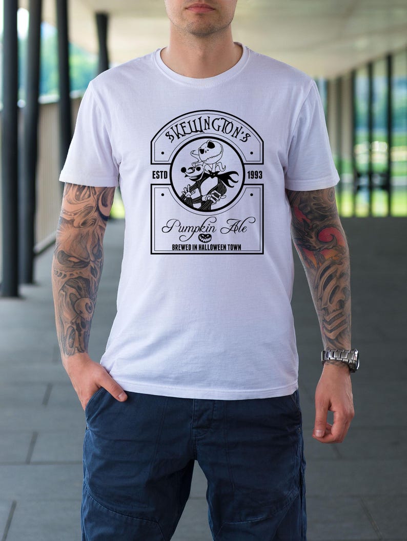 Puede incluir: Camiseta blanca con un dise&ntilde;o gr&aacute;fico en blanco y negro para "Skellington's Pumpkin Ale". El dise&ntilde;o incluye un emblema circular con un personaje esqueleto y el texto "Brewed in Halloween Town".