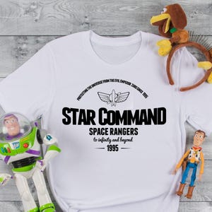 Op de afbeelding: Wit T-shirt met zwarte tekst: "STAR COMMAND SPACE RANGERS to infinity and beyond 1995". Het shirt heeft een ruimteschipafbeelding. Speelfiguren van Buzz Lightyear en Woody staan in de buurt van het shirt. De tekst luidt ook "PROTECTING THE UNIVERSE FROM THE EVIL EMPEROR ZURG SINCE 1995".