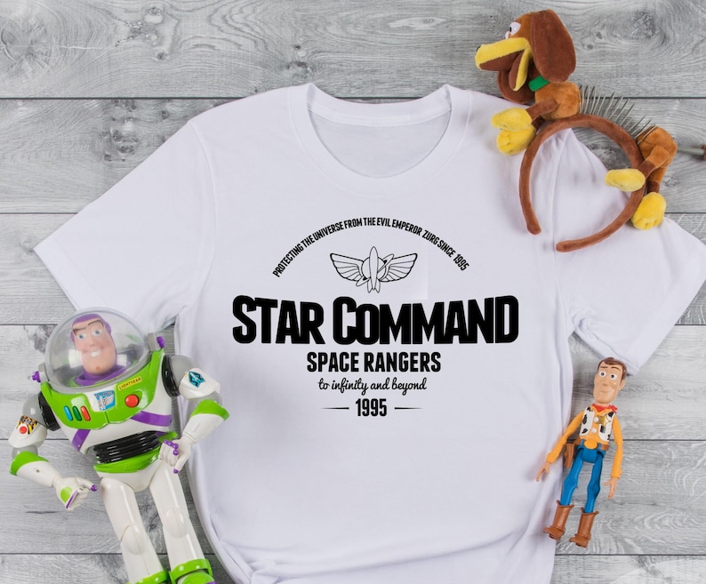 Op de afbeelding: Wit T-shirt met zwarte tekst: "STAR COMMAND SPACE RANGERS to infinity and beyond 1995". Het shirt heeft een ruimteschipafbeelding. Speelfiguren van Buzz Lightyear en Woody staan in de buurt van het shirt. De tekst luidt ook "PROTECTING THE UNIVERSE FROM THE EVIL EMPEROR ZURG SINCE 1995".