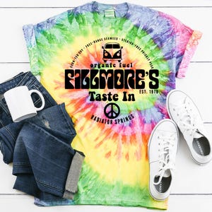May include: A tie-dye t-shirt with the text "Fillmore's Taste In" and a peace sign, paired with blue jeans, a white mug, and white sneakers. The shirt features a colorful swirl design with shades of yellow, green, blue, and pink.