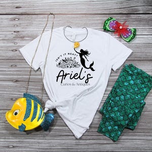 May include: White t-shirt with a mermaid graphic and the text "Isn't it neat? Ariel's Curios & Antiques." Also, a yellow and blue fish-shaped purse, green leggings with a scale pattern, and a decorative hair clip with a red bow.