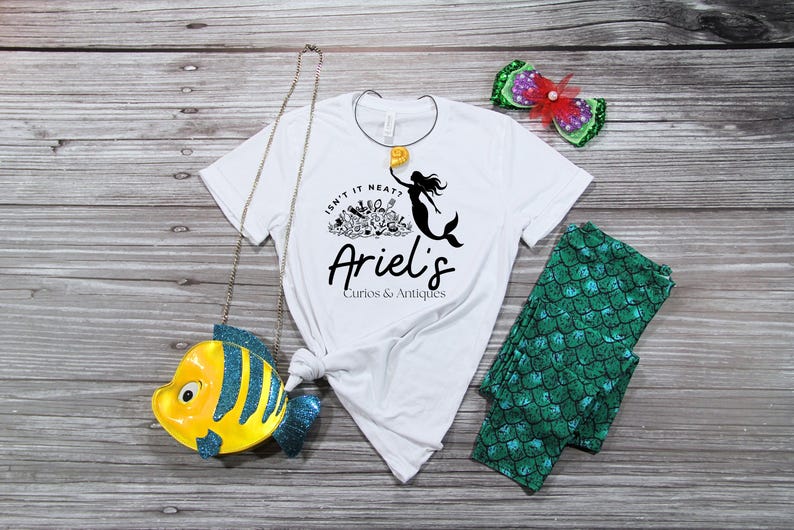 May include: White t-shirt with a mermaid graphic and the text "Isn't it neat? Ariel's Curios & Antiques." Also, a yellow and blue fish-shaped purse, green leggings with a scale pattern, and a decorative hair clip with a red bow.