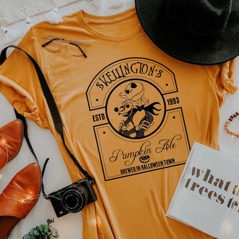 Puede incluir: Camiseta de color amarillo mostaza con un dise&ntilde;o negro de Jack Skellington y Zero de Pesadilla antes de Navidad, con el texto "SKELLINGTON'S PUMPKIN ALE BREWED IN HALLOWEEN TOWN". Tambi&eacute;n se ven un sombrero negro, una c&aacute;mara y un libro.