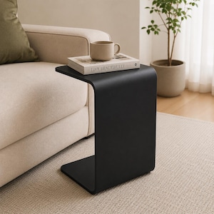 Puede incluir: Una mesa auxiliar negra en forma de C con un diseño curvo. Un libro con el texto "LIVE BEAUTIFULLY" y una taza beige están encima. La mesa está junto a un sofá beige y una planta en maceta.