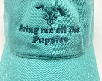 May include: A teal blue baseball cap with a cartoon dog graphic and the text "Bring me all the puppies" embroidered in dark green.