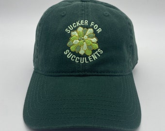 May include: A dark green baseball cap with the embroidered text "SUCKER FOR SUCCULENTS" above a green and gold succulent design. The cap has a curved brim and a classic six-panel design.