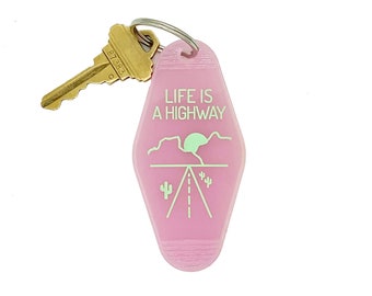 May include: Pink motel key chain with a gold key attached. The key chain has the text "Life is a Highway" and a graphic of a desert road with a sunset.