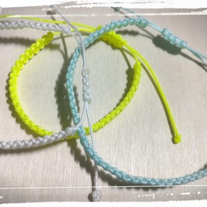 Op de afbeelding: Drie verstelbare koordarmbanden met een gevlochten ontwerp. Eén armband is wit, één is neongeel en één is lichtblauw. Elke armband is voorzien van een schuifknoopsluiting met bijpassende koorduiteinden voor een verstelbare pasvorm om de pols.