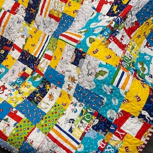 May include: A patchwork quilt with a vibrant design featuring Dr. Seuss characters and patterns. The quilt incorporates various fabrics in red, yellow, blue, and white, with striped and polka dot accents. The edges are finished with a red and white striped border.