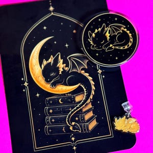 Peut inclure: Une impression rectangulaire noire représentant un dragon noir endormi sur un croissant de lune au-dessus d'une pile de livres, un support de téléphone circulaire noir avec un dragon endormi et une petite breloque dragon jaune.