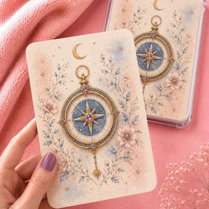 Peut inclure: Une carte rectangulaire présentant une illustration détaillée d'une boussole dorée au cadran bleu, entourée de motifs floraux rose et bleu doux et d'un croissant de lune. La boussole inclut les points cardinaux et des accents de pierres violettes.