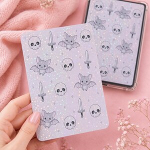 Peut inclure: Une planche d'autocollants rectangulaire et une coque de téléphone assortie présentant un motif répétitif de chauves-souris grises, de crânes blancs et d'épées argentées sur un fond lavande clair avec une finition pailletée holographique.
