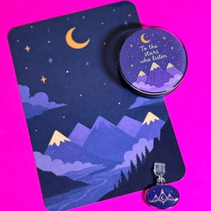 Peut inclure: Une carte rectangulaire illustrant un ciel nocturne avec un croissant de lune, des étoiles et des montagnes violettes, un support de téléphone circulaire avec le texte To the stars who listen, et une petite breloque ovale aux tons bleu et violet.