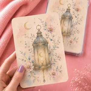 Peut inclure: Une carte rectangulaire présentant une illustration à l'aquarelle d'une lanterne ornée entourée de fleurs roses délicates, de feuillage bleu et d'un croissant de lune doré. La lanterne a une lueur douce et une petite breloque en cristal violet.