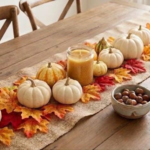 Peut inclure: Un chemin de table en toile de jute décoré de citrouilles blanches et orange, d'une bougie jaune dans un pot en verre, de feuilles d'automne artificielles rouges, orange et jaunes, et d'un bol en céramique rempli de glands et de noisettes.