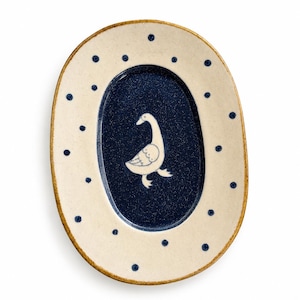 Pode incluir: Uma travessa oval de cerâmica com uma borda creme salpicada decorada com bolinhas azul-escuras. O centro da travessa é um oval azul-escuro profundo e sólido, que contém uma ilustração de linhas brancas de um ganso virado para a esquerda.