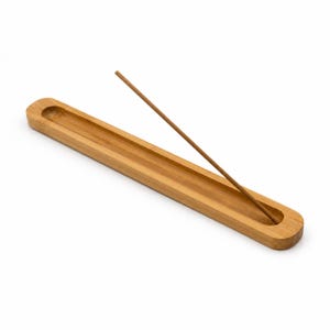 Peut inclure: Un porte-encens en bois brun clair, long et étroit, de forme rectangulaire arrondie et lisse, doté d'une rainure centrale. Un bâton d'encens brun, fin et unique, est placé en diagonale, reposant dans la rainure creusée du support.