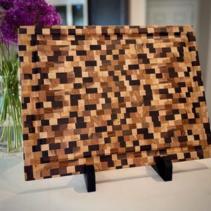 May include: A rectangular end-grain cutting board with a mosaic pattern of light and dark wood squares. The board is displayed on a black stand. A vase of purple flowers is visible in the background.