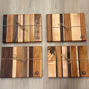 May include: Four rectangular wooden cutting boards featuring vertical stripes of varying light, medium, and dark brown wood tones. Each board is tied with a thin brown twine string and features a small black logo with the letters AD in the corner.