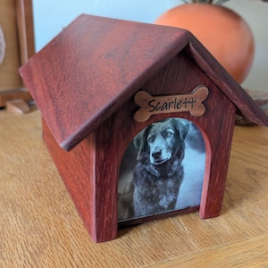 May include: A wooden dog house-shaped pet memorial urn with a photo of a dog inside. The roof and walls are a dark reddish-brown. A bone-shaped plaque above the entrance reads "Scarlett".